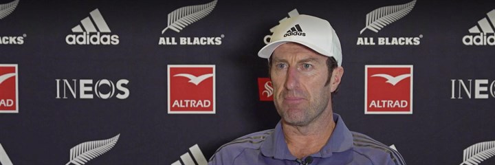 Dr Nic Gill, wearing a white cap and purple shirt, in front of a media background with the All Blacks logo